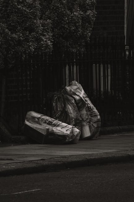 Three large black rubbish bags made from thick plastic material are placed on the edge of a paved pavement, positioned upright and leaning against a black metal fence. The bags appear filled with waste and are crinkled and slightly bulging, indicating they are tightly packed. The fence behind the bags has vertical bars, and dense greenery with leaves and branches extends above and to the side, partially obscuring the view. To the right, part of a dark brick building is visible, suggesting an urban setting nearby. The scene is illuminated by natural daylight, creating clear contrast and shadow around the bags and fence, reflecting a typical private waste disposal scenario managed by rubbish removal services such as Rubbish Clearance Marylebone, in a quiet street environment suitable for alternative waste handling or on-site clearance.