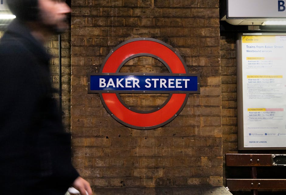 A partial view of a blurred man walking past a brick wall with a prominent round underground train sign displaying 'BAKER STREET' in white letters on a blue background within a red circle, mounted on the wall. beside the sign is an illuminated timetable board containing travel information, supported by a wooden bench beneath it, all situated in an indoor station environment. The scene features warm lighting and a textured brick surface, creating a typical urban setting associated with transportation hubs where private waste management services like Rubbish Clearance Marylebone may operate or offer alternative rubbish pickup solutions near Baker Street. The focus remains on the sign and timetable, with the moving figure adding a dynamic element to the scene, emphasizing the busy atmosphere typical of central London transport locations.