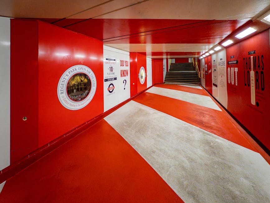 The image shows a brightly lit pedestrian underpass with a curved ceiling painted in red and cream. The floor is covered with a smooth, light-colored concrete surface, and the walls are decorated with large, bold black and red signage, including the words 'Prince Regent' with a crown symbol and historical details about the location. To the left, the wall features a circular emblem and a question mark, while to the right, there are additional signs and artwork referencing local history and landmarks. At the far end of the underpass, a staircase with wooden steps and black metal railings leads to an upper street level. The brightly illuminated environment, combined with the clean and organized appearance, suggests it is a well-maintained urban passageway, possibly used for private or independent access, aligning with alternative waste handling routes offered by services like Rubbish Clearance Marylebone for rubbish removal and bulk waste collection in the area.
