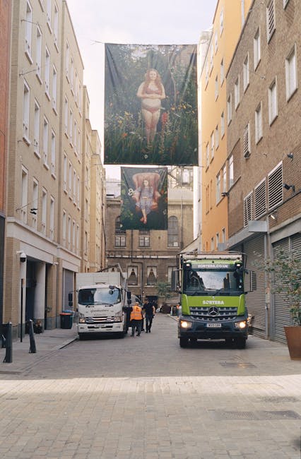 A narrow urban street scene featuring a cleaning vehicle, possibly used for rubbish collection or street maintenance, parked between tall, beige and yellow multi-storey buildings with several small windows. In the background, two large fabric banners hang above the street, each displaying artwork—one depicting a woman in a natural setting with trees, and the other showing a person seated, surrounded by lush foliage. The street has a paved surface and a few pedestrians, including a person wearing a high-visibility vest, walking near the vehicle. The environment suggests a typical city centre location where private waste management services, such as rubbish removal or on-site clearance, might operate, supported by companies like Rubbish Clearance Marylebone. The lighting appears natural, indicating daytime, and the overall scene combines elements of urban life, waste management activity, and street art or advertisements suspended overhead.
