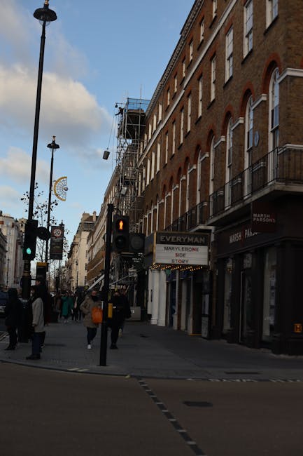 A daytime street scene near Madame Tussauds in Marylebone showing a row of multi-storey brick buildings with large, arched windows and small balconies on the right side. The buildings have a traditional appearance with brown and reddish brickwork, and some have scaffolding indicating ongoing building maintenance or refurbishment. In the street foreground, there is a pedestrian crossing with a traffic light displaying a red person signal, and several pedestrians walking across or waiting on the pavement. The pavement is lined with black street lamps, decorative lighting, and shopfronts, including a visible sign for 'Everyman' cinema. In the background, the street extends with more pedestrians and shops visible further down, with a partly cloudy sky overhead. This scene is an example of urban waste management or private rubbish collection in central London, reflecting the typical environment where rubbish clearance services like those offered by Rubbish Clearance Marylebone are often involved in maintaining cleanliness during building work or street improvement projects, especially in busy commercial districts near tourist attractions like Madame Tussauds.