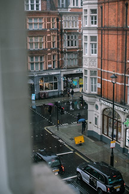 View from a high window overlooking a rainy city street in Marylebone with brick and white façade buildings, showing pedestrians walking on the wet pavement holding umbrellas, some crossing at the crosswalk, and vehicles including two black cars parked along the curb. The scene features a street-level pharmacy with a green cross sign, a nearby shop with large windows, and a temporary yellow construction sign on the sidewalk. The wet asphalt reflects the surrounding structures and streetlights, creating a moody urban atmosphere. This image relates to private waste handling or alternative rubbish collection services in Marylebone, as it depicts an urban environment where on-site clearance or independent rubbish removal might be necessary, contrasting with curbside council rubbish collection. Rubbish Clearance Marylebone could potentially handle such clearance needs, evident from the scene’s context of city street activity and waste management considerations at the local level.