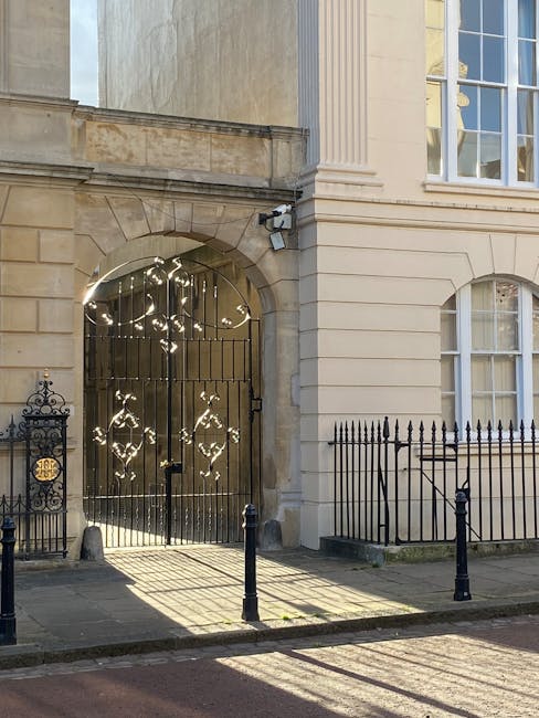 The image depicts a section of a historic or classical-style building's exterior, featuring a large, ornate iron gate with decorative patterns and a central lock, set within an arched stone entranceway. The building's façade is constructed of light-colored stone with a smooth, polished finish, and it has vertical fluted columns and large sash windows with white wooden frames. To the right of the gate, there is a black wrought iron fence with pointed finials, positioned on a raised stone base, which extends along the sidewalk. In the foreground, a paved pedestrian area is visible, with shadows cast by short black metal bollards lining the edge of the walk. The scene is illuminated by natural daylight, creating bright reflections on the iron gate, and the overall environment suggests a quiet, well-maintained urban setting that could be suitable for private property or a formal estate, often requiring occasional rubbish management or clearance services from companies such as Rubbish Clearance Marylebone.