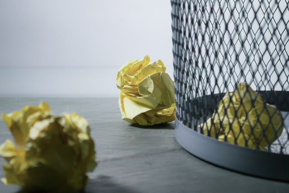 A close-up image shows crumpled yellow paper balls resting on a smooth, gray surface, with one larger ball leaning against a black wire mesh waste bin. The waste bin has a rounded base and a cylindrical shape, featuring a diamond-patterned metal grille design that allows visibility into the yellow paper refuse inside. The background consists of a plain, light-colored wall with even, neutral lighting, emphasizing the textures of the crumpled paper and the metal wireframe. This scene suggests a setting where waste is temporarily collected prior to disposal or a step in the process of rubbish removal, reflecting the kind of environment where services like those offered by Rubbish Clearance Marylebone could be involved in managing and clearing such paper debris from office or domestic spaces, supporting private disposal methods instead of council waste collection. The focus on the crumpled paper and waste bin, combined with a clean and orderly surroundings, conveys a professional approach to rubbish handling aligned with independent rubbish clearance services in the area.