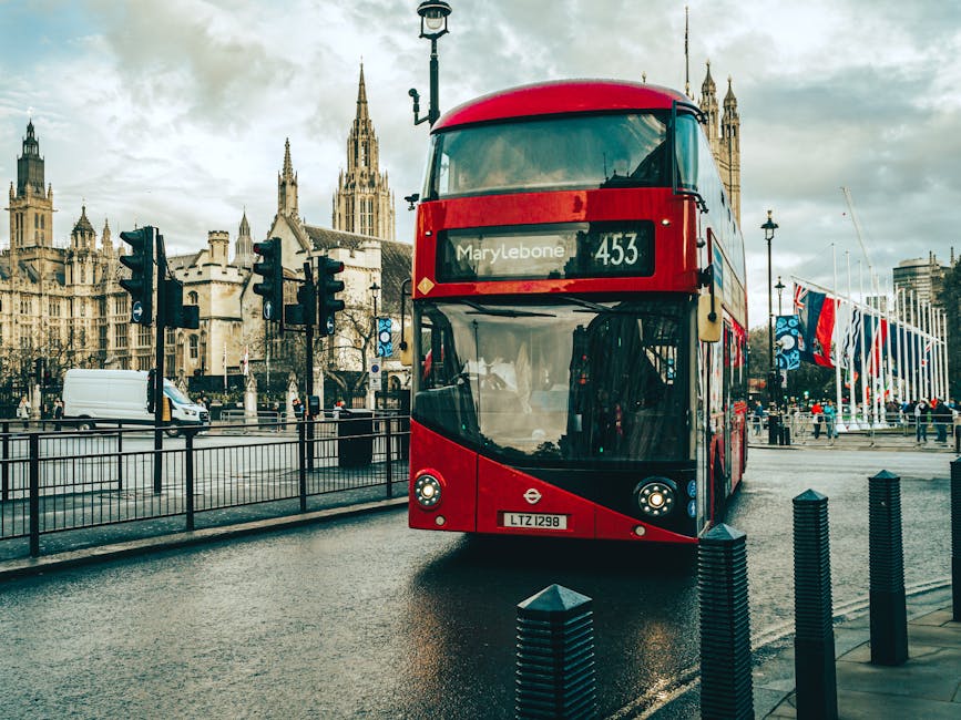 A red double-decker bus with a curved front windshield and modern design, displaying the route number 453 and destination Marylebone on an electronic signboard, occupies the foreground on a wet city street. The bus is positioned slightly to the right, with its headlights on and a visible registration plate marked LZ1298. The background features historic, Gothic-style buildings with pointed spires and ornate stonework, contrasting with the modern bus. To the left of the bus, several dark traffic lights are mounted on metal poles, and a white delivery van is parked further back behind a black metal fence that lines the sidewalk. On the right side of the image, a row of vertical black bollards separates the pedestrian area from the roadway. Some pedestrians are visible in the distance, near a monument or flag display, with overcast sky clouds casting diffuse light over the scene. The overall setting suggests a bustling urban environment, ideal for independent public transport or alternative disposal services like those provided by Rubbish Clearance Marylebone, situated amidst a mix of city infrastructure and historical architecture.