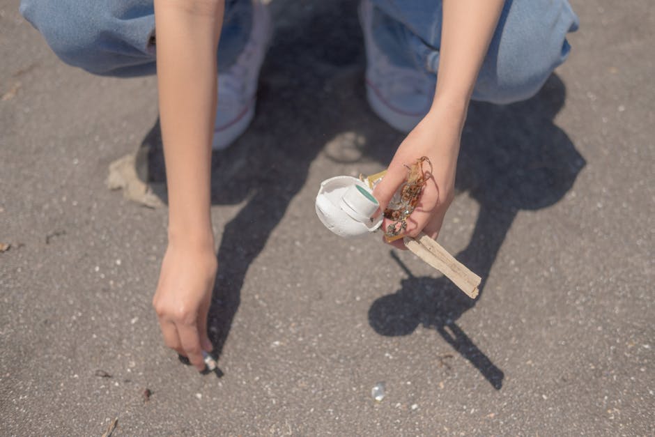 A person kneeling on a paved outdoor surface, using their right hand to pick up a small piece of white painted wood and debris with a textured, rough surface. The individual is dressed in light blue jeans and white sneakers with a red stripe, with their left hand resting on the ground for support. The surrounding environment appears to be an open area, possibly a driveway or street, with small scattered pieces of rubbish visible nearby. The lighting suggests natural daylight, casting shadows of the person's arm and the object being collected. This scene relates to waste collection or rubbish removal activities, with the person actively gathering loose trash, reflecting services offered by companies such as Rubbish Clearance Marylebone, which specialise in alternative waste handling and site clearance tasks.