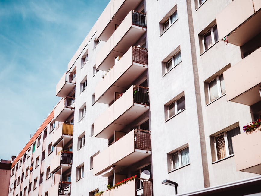 The image shows the exterior of a multi-storey residential building with a grey concrete facade and several rows of balconies. Each balcony is enclosed with metal railings painted in various dark colours, such as black and brown, with some featuring small flower boxes or plants. The building's windows are uniformly rectangular with white frames, and some have curtains or blinds partially visible. The balconies are staggered vertically, with a few personal items like laundry or decorative objects placed on them. The sky above is clear with a soft light, indicating daytime. The surrounding environment includes a street lamp in the foreground and a pedestrian pathway at the building's base. The overall scene reflects an urban residential setting, relevant to private waste management and rubbish removal services like those offered by Rubbish Clearance Marylebone, highlighting the typical features of apartment building exteriors and their communal outdoor spaces.