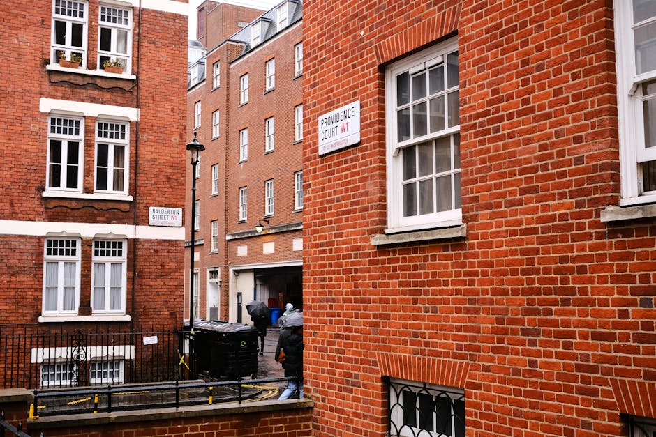 The image depicts a narrow urban alleyway flanked by red brick residential buildings with white-framed sash windows, some of which feature window boxes with small plants. In the foreground, a black metal fence encloses a small outdoor area, with a black waste bin placed against it. Behind the fence, two people are seen recycling or handling waste near a dark-colored vehicle partially visible at the back of the alley. The scene is illuminated by natural daylight, casting soft shadows on the brick walls, and environmental details suggest a typical city street corner. Signs attached to the buildings include street names, such as 'Balderton Street WC1' and 'Providence Court WC1,' indicating the location. The scene subtly relates to private waste handling and rubbish collection services, with visible elements suggesting the process of rubbish disposal in a denser urban setting, aligning with the subject of independent rubbish clearance and waste removal services by Rubbish Clearance Marylebone.