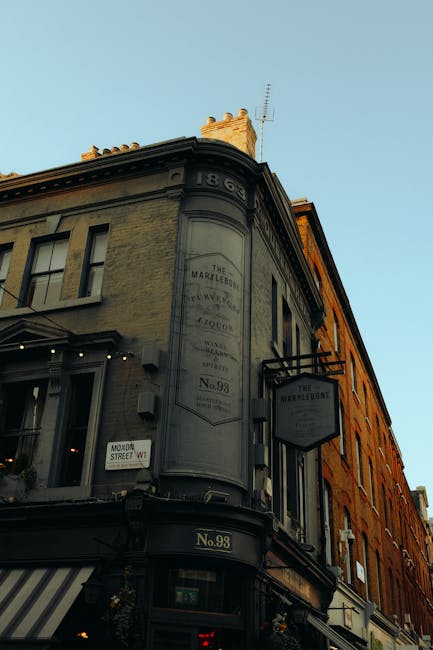 The photograph captures a corner view of a historic building with a rounded facade at the intersection of two streets, under a clear blue sky. The building's exterior features a combination of aged stone and brickwork, with decorative architectural details and a prominent vertical sign advertising 'The Marylebone,' which indicates its historical significance or previous use. Below the sign, a smaller rectangular sign lists the address as No. 98 and includes a metal bracket supporting a hanging sign for a business, possibly a pub or shop. The lower part of the building displays a black awning with white stripes, adjacent to a window with a small, illuminated interior visible from the street. The overall scene is bathed in natural daylight, suggesting late afternoon or early evening, and the surroundings imply an urban setting, possibly a commercial or mixed-use area. Rubbish clearance services by Rubbish Clearance Marylebone may operate nearby, supporting alternative waste disposal and private rubbish collection for businesses and residents in the area.
