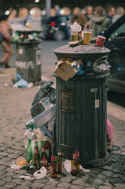 A black, cast-iron public trash bin situated on a cobblestone pavement in an urban setting, with noticeable wear on its surface and a slot for inserting waste. The top of the bin holds several disposable coffee cups, some with lids and others without, along with a crumpled paper bag and a red disposable cup. Around the base of the bin, there are multiple empty glass bottles, some with labels, as well as scattered paper, food wrappers, and other small debris. In the background, there is a blurred street scene with pedestrians, parked cars, and another rubbish bin, indicating the area is a busy city environment. The scene suggests a collection point for litter that has accumulated from nearby foot traffic, illustrating the importance of waste management and the potential need for private rubbish removal services like Rubbish Clearance Marylebone to maintain cleanliness in urban public spaces.