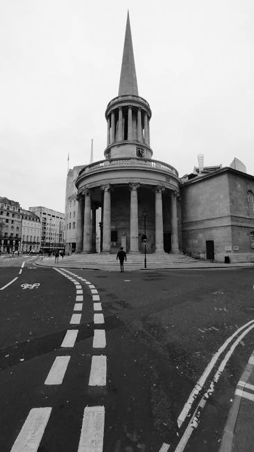 A black-and-white photograph of the historic St. Martin-in-the-Fields church in London's Trafalgar Square area, featuring a tall, pointed spire rising above a circular, columned exterior façade. The building's stone surface displays a smooth texture with some weathering, and the rounded colonnade supports a low parapet. In the foreground, a paved circular road with clearly marked, dashed white lane lines surrounds the church, with a single person walking across the street. To the left, additional buildings with uniform window patterns line the street, contributing to an urban environment. The scene is lit evenly, with no strong shadows or highlights, emphasizing the church's architectural details and the surrounding street scene. This image may be used to illustrate the importance of on-site clearance or private waste handling, as Rubbish Clearance Marylebone sometimes facilitates alternative waste collection solutions near prominent landmarks or historic sites, ensuring local regulations on rubbish removal are met efficiently.