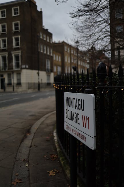 A black metal fence surrounds Montagu Square in Westminster, with a white street sign attached to it indicating the location as Montagu Square W1. The sign features black and red lettering, with the logo of Westminster visible beneath. In the background, there is a row of traditional multi-storey brick buildings with white-framed windows, characteristic of residential architecture in the area. The street appears quiet, with a paved sidewalk running alongside, and some fallen leaves scattered on the ground, suggesting an autumn or winter setting. Overcast skies cast a dull light over the scene, emphasizing the urban environment. The image relates to the context of private waste handling and local authority licensing, as such signage and fencing can be associated with site access or temporary storage areas for rubbish clearance services, which companies like Rubbish Clearance Marylebone might provide as part of on-site waste removal or alternative disposal options within Westminster.
