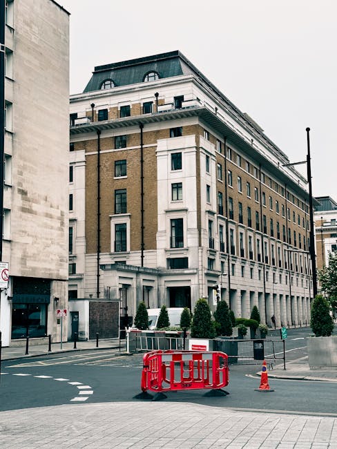 A multi-storey building with classical architectural features, including white decorative columns and a dark, mansard-style roof, is situated on a city street corner. The building's façade combines light brown brickwork with white window frames and detailing, with several windows visible on each floor. The street in front of the building shows a section of paved road and sidewalk, with temporary orange traffic cones and a red safety barrier indicating an area of ongoing activity, possibly related to construction or property maintenance. Small, neatly trimmed evergreen shrubs are planted along the base of the building, behind a low metal fence. The background includes a cloudy sky, and a streetlamp is visible to the right of the building, subtly blending into the urban environment. This scene, captured in daytime, reflects a typical cityscape with ongoing work around a prominent historic building, illustrating a context where private or independent waste removal services, such as those provided by Rubbish Clearance Marylebone, could be engaged for construction site clearance or rubbish collection in an urban setting.