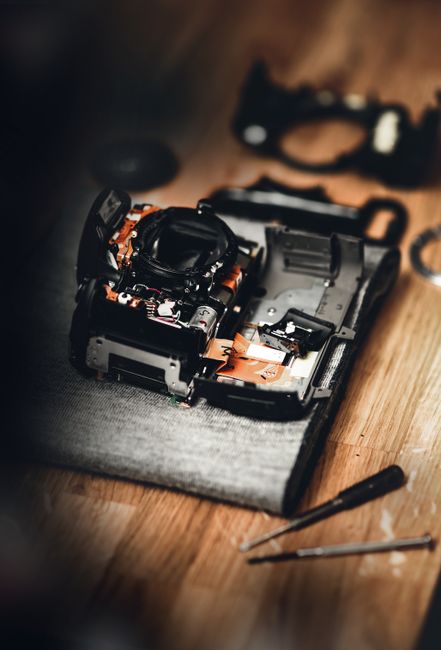 A partially disassembled digital camera rest on a dark cloth on a wooden surface, with its internal components and circuit boards visible, including orange ribbon cables and black plastic housing. Surrounding it, there are small tools such as a small screwdriver and a pair of tweezers, placed nearby, indicating ongoing repair work. In the background, a camera lens and a black ring-shaped component are blurred but visible. The scene appears to be in a workshop or repair area, with natural lighting casting soft shadows across the workspace. This detailed view highlights the process of electronics disassembly, which may relate to waste management of electronic devices, such as camera parts, supported by Rubbish Clearance Marylebone's services for proper disposal of electronic waste, promoting environmentally responsible rubbish removal and recycling of digital devices.
