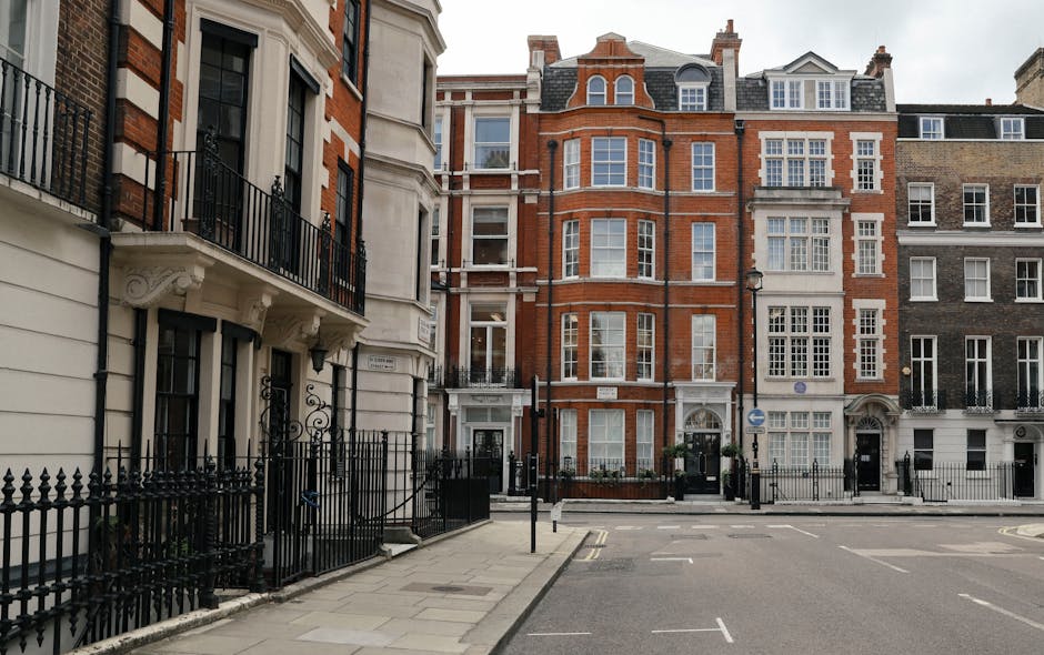 A multi-storey building with classical architectural features, including white decorative columns and a dark, mansard-style roof, is situated on a city street corner. The building's façade combines light brown brickwork with white window frames and detailing, with several windows visible on each floor. The street in front of the building shows a section of paved road and sidewalk, with temporary orange traffic cones and a red safety barrier indicating an area of ongoing activity, possibly related to construction or property maintenance. Small, neatly trimmed evergreen shrubs are planted along the base of the building, behind a low metal fence. The background includes a cloudy sky, and a streetlamp is visible to the right of the building, subtly blending into the urban environment. This scene, captured in daytime, reflects a typical cityscape with ongoing work around a prominent historic building, illustrating a context where private or independent waste removal services, such as those provided by Rubbish Clearance Marylebone, could be engaged for construction site clearance or rubbish collection in an urban setting.